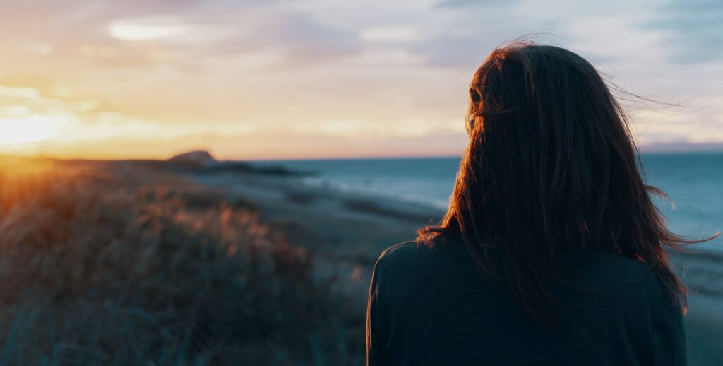 a woman is standing on the beach at sunset looking out at the water to represent mental health services in newport beach. 